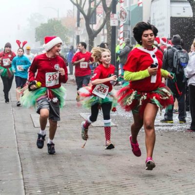 Excited runners make their way toward the finish line of the Spirit of Giving 5K Run as snow falls from above and spectators cheer them on.