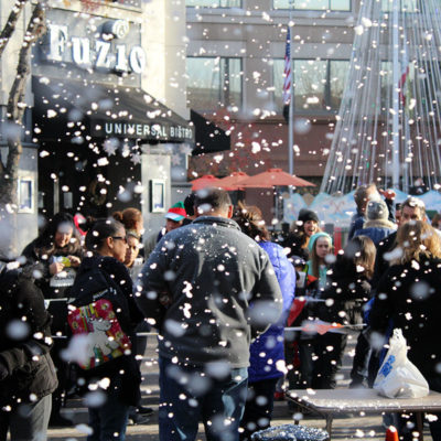Spectators and runners enjoy the festive scene after the finish line of the Spirit of Giving 5K Run/Walk. The race features artificial snow coming from the Downtown Modesto buildings and falling on the race festival below.